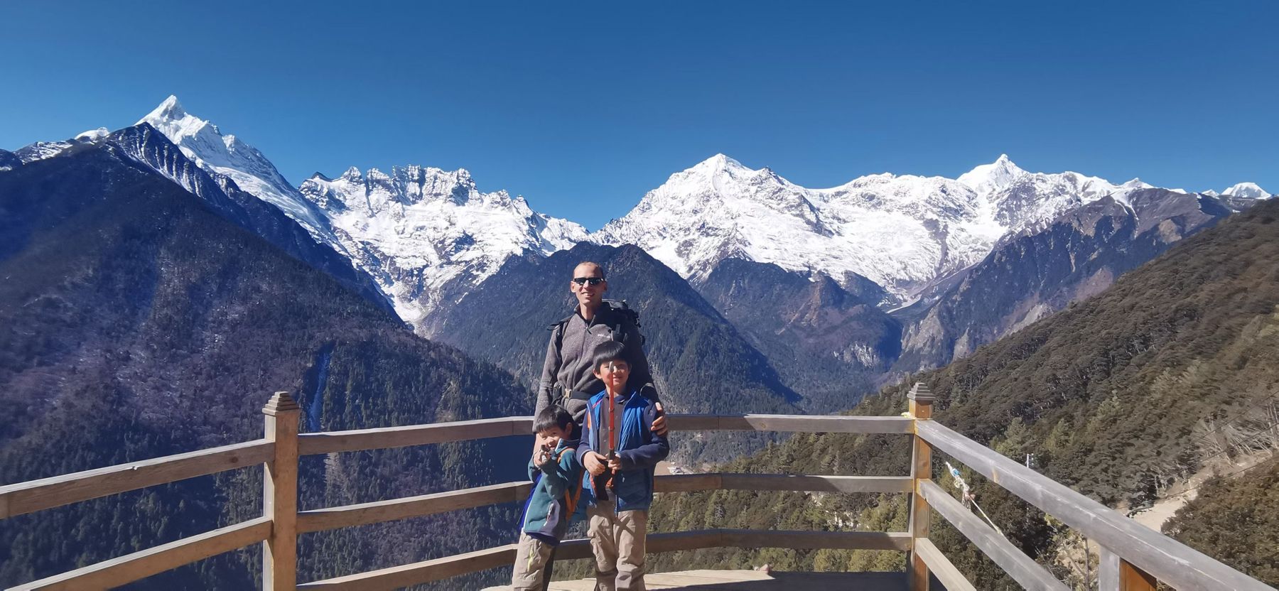 Un homme et un garçon à une balustrade en bois sur un col, face à une chaîne de sommets enneigés