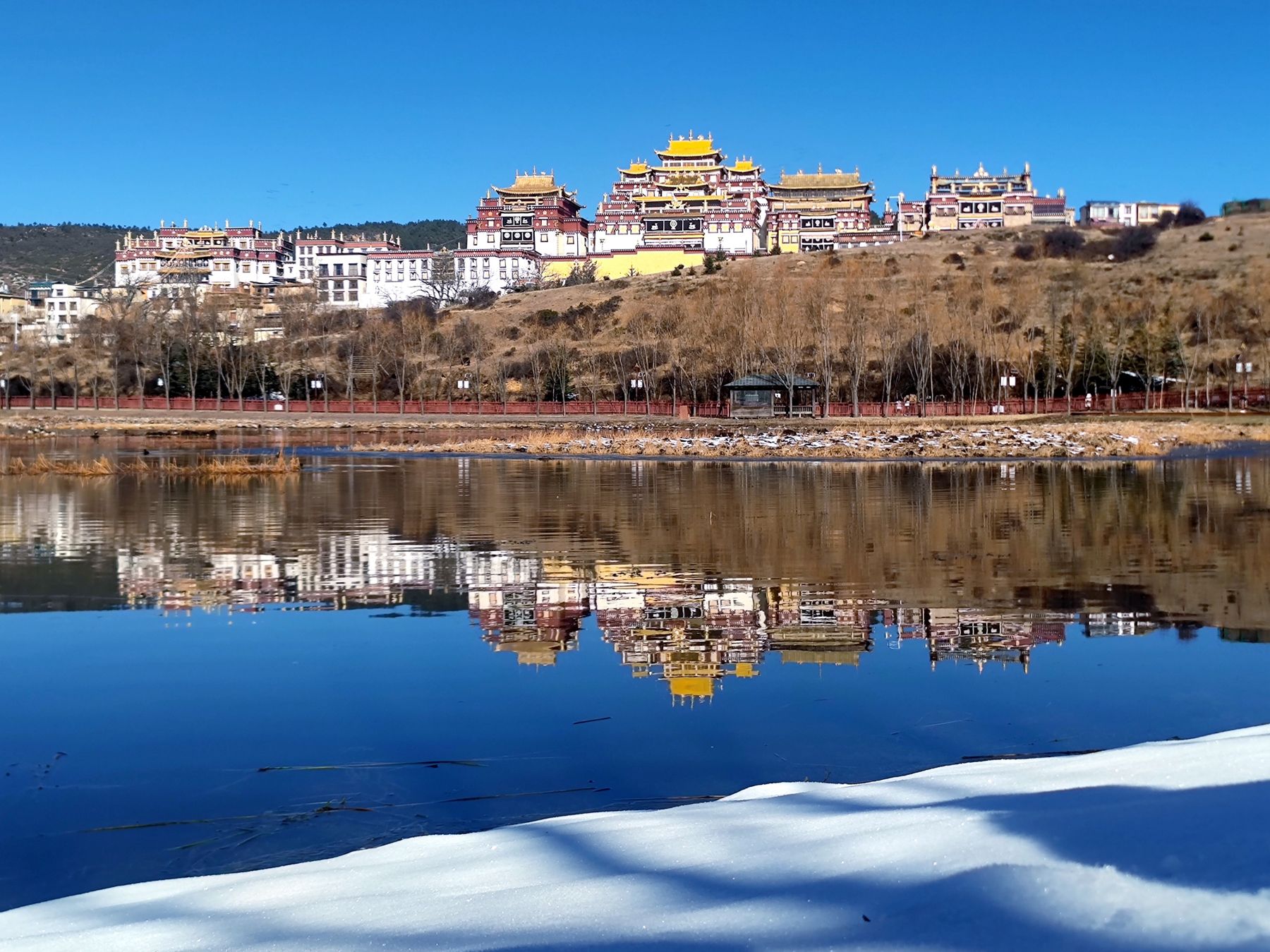 Un monastère aux toits dorés se reflétant dans un lac, avec de la neige au premier plan